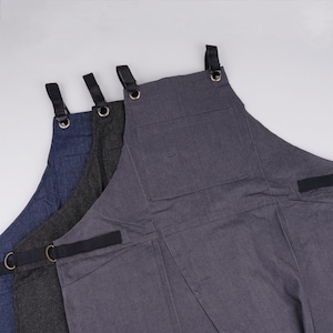 May include: Three denim aprons in different shades of blue, black, and gray. Each apron has adjustable straps and a large pocket on the front.