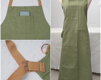 May include: Olive green apron with brown straps and gold-colored metal rings. The apron has a large front pocket and a smaller pocket on the side. The straps are adjustable. The apron is made of a durable fabric.