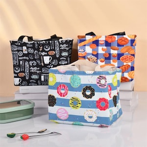 Op de afbeelding: Drie geïsoleerde lunchtassen met verschillende patronen met een voedselthema. Eén tas heeft donuts op blauw-witte strepen, een andere heeft een zwarte achtergrond met ontbijtproducten en de derde heeft een blauw-wit geblokt patroon met brood.