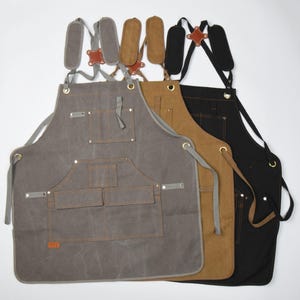 May include: Three aprons in gray, brown, and black, each with adjustable straps and multiple pockets. The aprons have leather accents and metal hardware. The gray apron is on the left, the brown in the middle, and the black on the right.