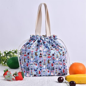 Pode incluir: Uma lancheira azul clara com cordão e alça creme. A bolsa apresenta um padrão repetido de personagens de Alice no País das Maravilhas e elementos florais. A bolsa é mostrada com frutas frescas.