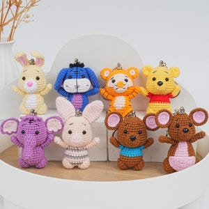 May include: A collection of handmade crochet keychains featuring various cartoon characters. The keychains include a yellow bunny, a blue donkey, an orange tiger, a yellow bear, a purple elephant, a pink bunny, a brown bear, and a brown mouse.
