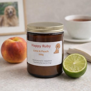 Könnte beinhalten: Duftkerze in einem braunen Glasbehälter mit goldenem Deckel, beschriftet mit "Happy Ruby Lime & Peach 200g". Das Etikett zeigt eine Hundeillustration. Eine Pfirsichhälfte und eine Limettenhälfte liegen neben der Kerze.