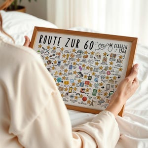 May include: A framed print with the text "ROUTE ZUR 60" and "GEBOREN 1966" in black. The print features numerous small illustrations and stars, all within a light brown wooden frame. The print is held by a person.