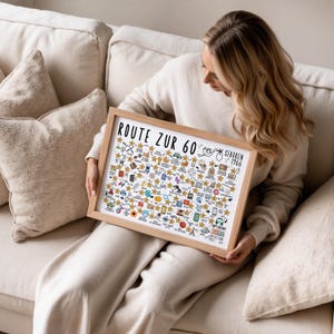 May include: A framed print with the text "ROUTE ZUR 60" and "GEBOREN 1966" in black. The print features numerous small illustrations and gold stars. The frame is made of light-colored wood. The person is sitting on a beige couch.