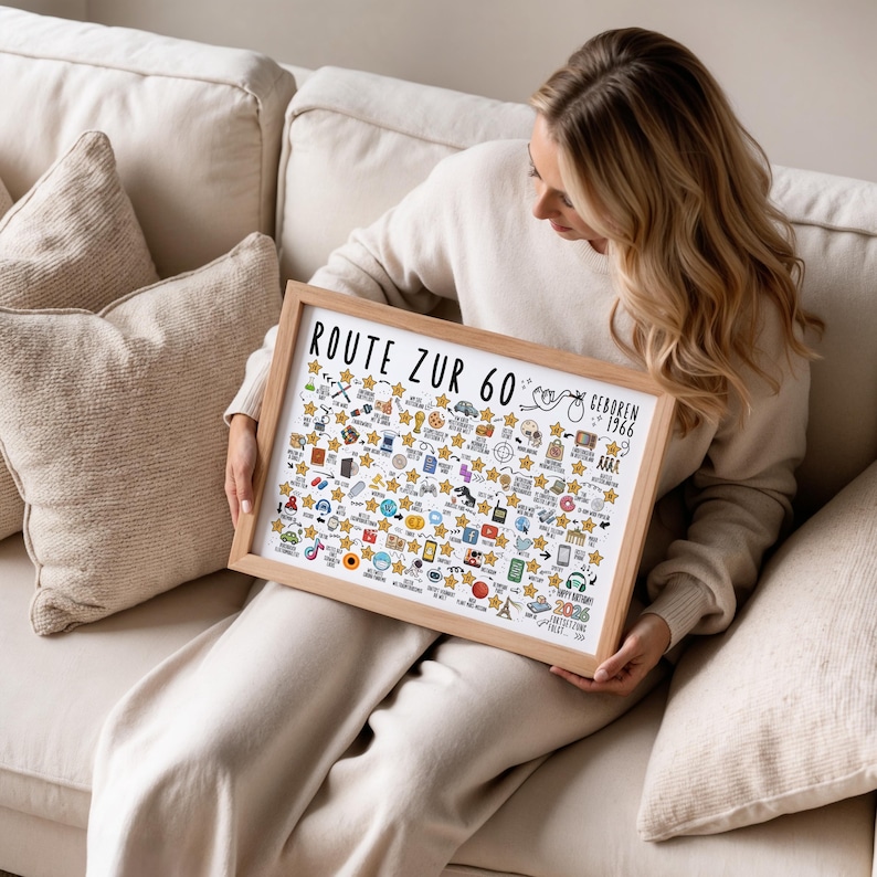 May include: A framed print with the text "ROUTE ZUR 60" and "GEBOREN 1966" in black. The print features numerous small illustrations and gold stars. The frame is made of light-colored wood. The person is sitting on a beige couch.