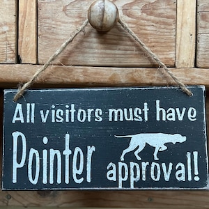 May include: Black rectangular wooden sign with white text: "All visitors must have Pointer approval!" A white silhouette of a dog is on the right. The sign hangs from a twine string.