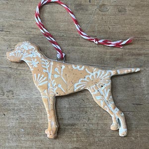 May include: A tan ceramic dog ornament with white floral patterns. It features a red and white string for hanging. The dog is depicted standing, with its tail extended. The ornament is approximately 15 cm long.