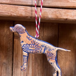 May include: A ceramic dog ornament with a tan base and blue floral patterns. The dog is suspended by a red and white striped string. The ornament is a decorative item.