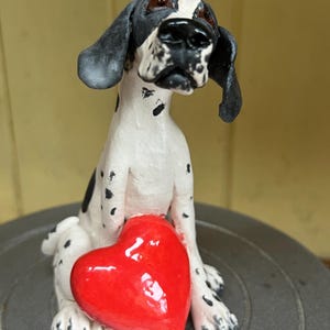 May include: A ceramic dog figurine with black and white markings, holding a red heart. The dog has a sad expression and floppy ears. The heart is a glossy red. The figurine is a decorative item.