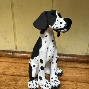 May include: A ceramic dog figurine, featuring a black and white spotted design. The dog is depicted in a seated pose. The figurine is placed on a wooden surface.
