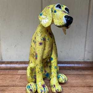 May include: A ceramic dog ornament in a seated pose. The dog is painted yellow with speckled accents of black, blue, and purple. The dog has black eyes and a black nose. The ornament is on a wooden surface.