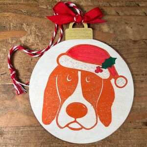 May include: White wooden Christmas bauble featuring a brown and orange dog wearing a red and white Santa hat. The bauble has a red and white string for hanging.