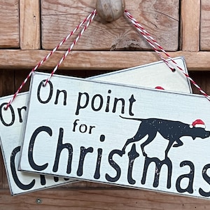 May include: Two white wooden signs with black lettering and a black silhouette of a dog wearing a red Santa hat. The top sign reads "On point for Christmas". The bottom sign reads "On Christmas".