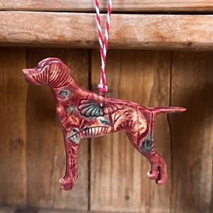 May include: A ceramic dog ornament in shades of red, with teal and gold accents. The dog is suspended by a red and white string. The ornament has a detailed, textured surface and is likely handmade. The dog is a Pointer breed.
