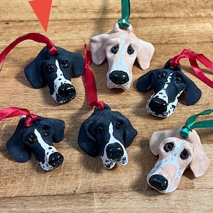 May include: Handmade ceramic dog head ornaments with red and green ribbon hangers. The ornaments feature black and white or tan and white spotted dog faces. These decorative ornaments are perfect for dog lovers and holiday decor.
