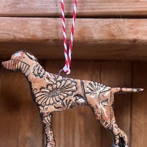 May include: A ceramic dog ornament with a brown base colour and black floral and geometric patterns. The ornament is suspended by a red and white striped string. The dog is in a standing position.