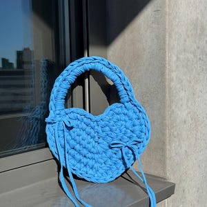 May include: A sky blue, heart-shaped crochet handbag with a braided handle and long, dangling ties. The bag is made of thick, textured yarn and sits on a windowsill. The bag's shape and color give it a playful, summery feel.