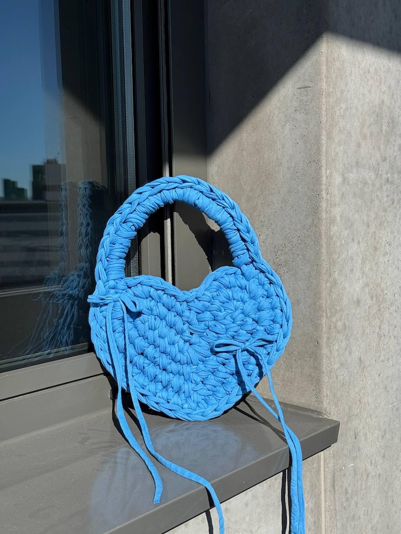 May include: A sky blue, heart-shaped crochet handbag with a braided handle and long, dangling ties. The bag is made of thick, textured yarn and sits on a windowsill. The bag's shape and color give it a playful, summery feel.