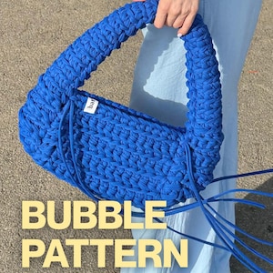 Pode incluir: Uma bolsa de mão em azul royal vibrante feita de tecido trançado grosso com formato de meia-lua e uma alça única integrada. Uma pequena etiqueta branca com texto preto está fixada na lateral. Longos cordões azuis pendem da base da alça.