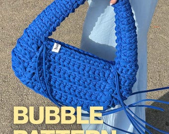 May include: A vibrant royal blue handbag crafted from thick, braided fabric with a crescent shape and a single integrated handle. A small white label with black text is attached to the side. Long blue cord accents hang from the base of the handle.