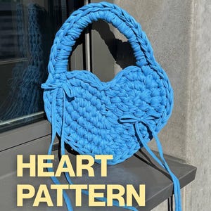 May include: A bright blue heart-shaped handbag crafted from thick, braided fabric yarn. The bag features a sturdy braided handle and two decorative fabric ties on the sides. The text "HEART PATTERN" is displayed in yellow letters at the bottom.