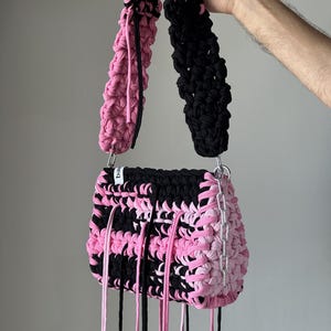 Może przedstawiać: Ręcznie wykonana szydełkowa torebka z wzorem typu color-block z czarnej i jasnor&oacute;żowej wł&oacute;czki. Torebka posiada gruby, teksturowany pasek na ramię, srebrny metalowy łańcuszek z boku oraz długie, zwisające sznurki z tkaniny.