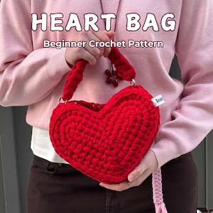 May include: A handmade red crochet heart bag with the text "HEART BAG" and "Beginner Crochet Pattern". The bag has a braided handle and a long strap. The bag is held by a person wearing a pink sweater.