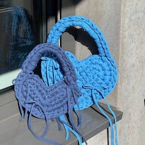 May include: Two handmade, heart-shaped bags in shades of blue. The bags are made of a thick, knitted material with braided handles and long, dangling ties. One bag is a lighter shade of blue, while the other is a darker, navy blue.
