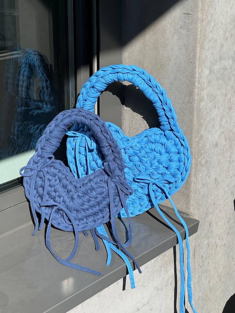 May include: Two handmade, heart-shaped bags in shades of blue. The bags are made of a thick, knitted material with braided handles and long, dangling ties. One bag is a lighter shade of blue, while the other is a darker, navy blue.