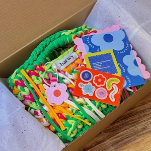 K&ouml;nnte beinhalten: Eine farbenfrohe, handgefertigte Tragetasche in einem Karton. Die Tasche besteht aus gr&uuml;nem, rosa, gelbem und wei&szlig;em Stoff. Die Box enth&auml;lt au&szlig;erdem Aufkleber, Anh&auml;nger und Karten mit floralen und textlichen Designs. Der Markenname "barwy" ist auf einem Anh&auml;nger zu sehen.