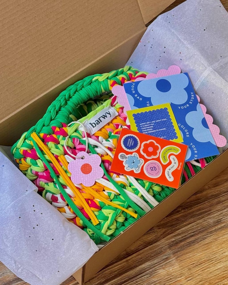 K&ouml;nnte beinhalten: Eine farbenfrohe, handgefertigte Tragetasche in einem Karton. Die Tasche besteht aus gr&uuml;nem, rosa, gelbem und wei&szlig;em Stoff. Die Box enth&auml;lt au&szlig;erdem Aufkleber, Anh&auml;nger und Karten mit floralen und textlichen Designs. Der Markenname "barwy" ist auf einem Anh&auml;nger zu sehen.
