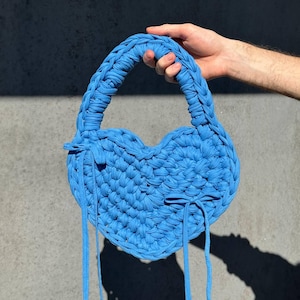 May include: A heart-shaped, sky blue handbag with a braided handle and long, thin straps. The bag is made of a thick, textured material, and is held up by a person's hand. The bag's shadow is cast on a gray surface.