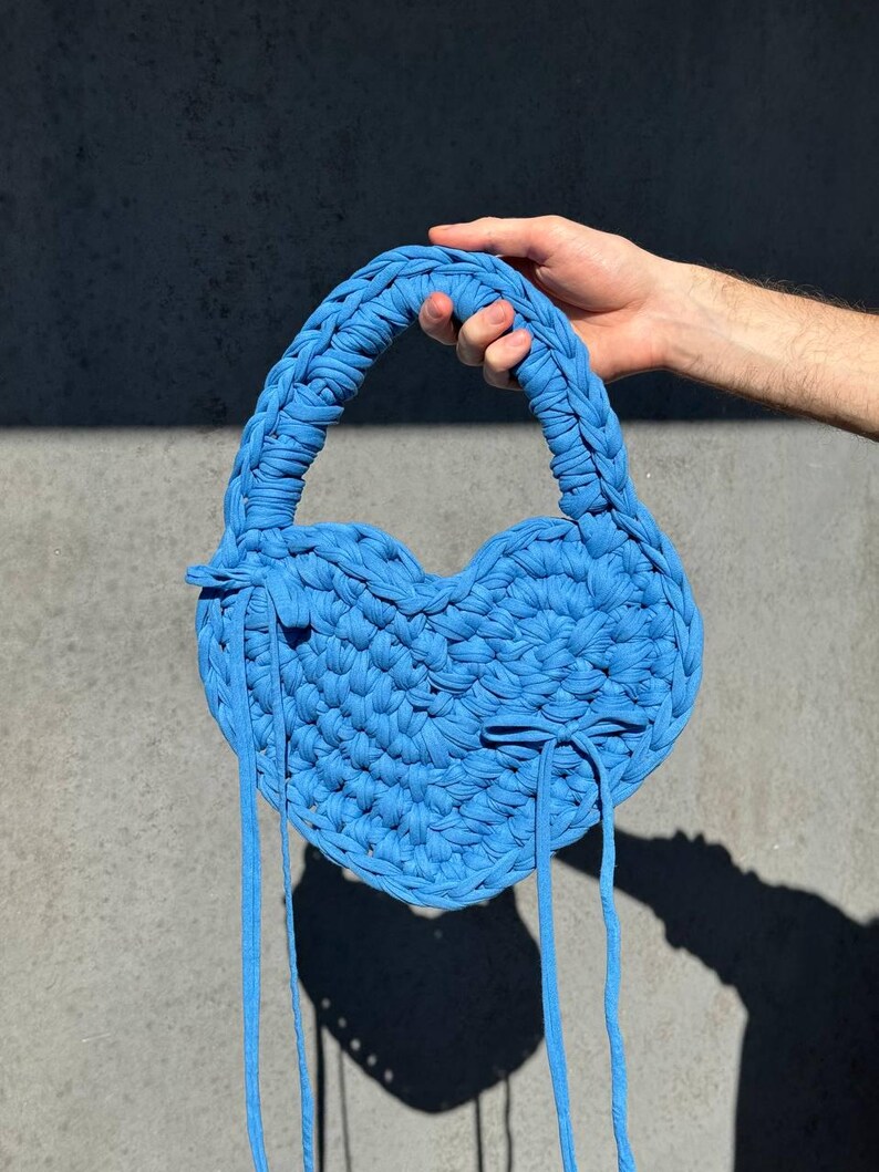 May include: A heart-shaped, sky blue handbag with a braided handle and long, thin straps. The bag is made of a thick, textured material, and is held up by a person's hand. The bag's shadow is cast on a gray surface.