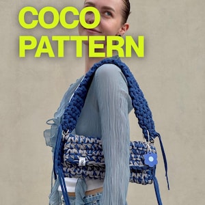 Può includere: Una borsa a tracolla rettangolare all'uncinetto con un motivo in filato mélange blu e grigio e una spessa tracolla intrecciata blu. La borsa include un piccolo ciondolo a forma di fiore blu e un'etichetta bianca con testo. Il testo COCO PATTERN appare in giallo brillante.