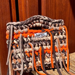 K&ouml;nnte beinhalten: Geh&auml;kelte Tragetasche in Braun-, Beige- und Oranget&ouml;nen. Die Tasche hat einen geflochtenen Griff und dekorative Fransen. Das Etikett auf der Tasche lautet "barwy".
