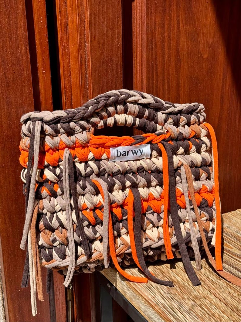 K&ouml;nnte beinhalten: Geh&auml;kelte Tragetasche in Braun-, Beige- und Oranget&ouml;nen. Die Tasche hat einen geflochtenen Griff und dekorative Fransen. Das Etikett auf der Tasche lautet "barwy".