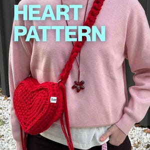 Puede incluir: Un bolso de ganchillo rojo brillante en forma de corazón con una correa trenzada gruesa y una pequeña etiqueta blanca con la palabra bar. El texto HEART PATTERN está superpuesto en azul claro. Un cordón oscuro con un colgante de flor roja cuelga.