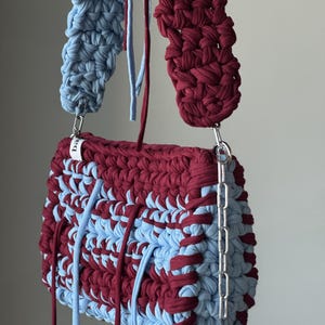 Może przedstawiać: Ręcznie wykonana szydełkowa torebka z wzorem typu color-block z bordowej i jasnoniebieskiej wł&oacute;czki. Torebka posiada gruby, pasujący szydełkowy pasek na ramię, srebrne metalowe okucia oraz długie, zwisające sznurki z tkaniny.