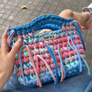 K&ouml;nnte beinhalten: Handgefertigte, farbenfrohe H&auml;keltasche mit blauem Griff und Boden sowie einer Mischung aus rosa, blauem und mintgr&uuml;nem Garn. Die Tasche hat lange, baumelnde Fransen in passenden Farben und ein Etikett mit der Aufschrift "barwy".