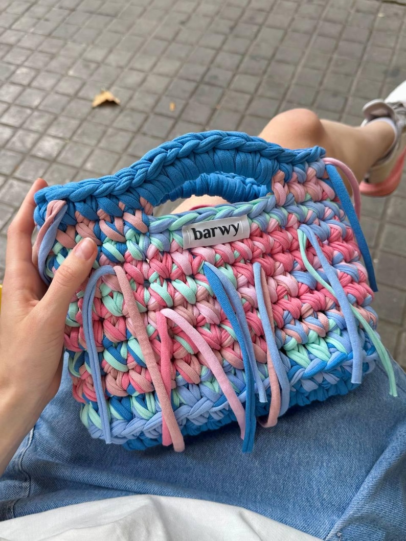 K&ouml;nnte beinhalten: Handgefertigte, farbenfrohe H&auml;keltasche mit blauem Griff und Boden sowie einer Mischung aus rosa, blauem und mintgr&uuml;nem Garn. Die Tasche hat lange, baumelnde Fransen in passenden Farben und ein Etikett mit der Aufschrift "barwy".
