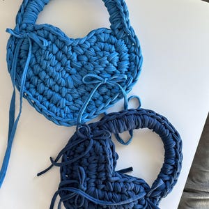 May include: Two heart-shaped, handmade bags in shades of blue. The bags are crafted from a thick, textured material, with a braided handle and decorative ties. The bags are a unique and stylish accessory.