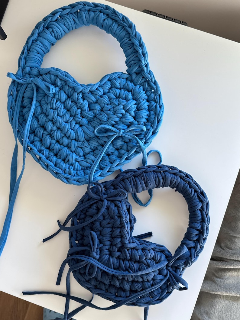 May include: Two heart-shaped, handmade bags in shades of blue. The bags are crafted from a thick, textured material, with a braided handle and decorative ties. The bags are a unique and stylish accessory.