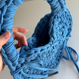 May include: A handmade, sky blue knitted handbag with a rounded handle. The bag is constructed from thick, braided fabric, and features a drawstring closure. The overall design is textured and soft, with a casual, yet stylish aesthetic.