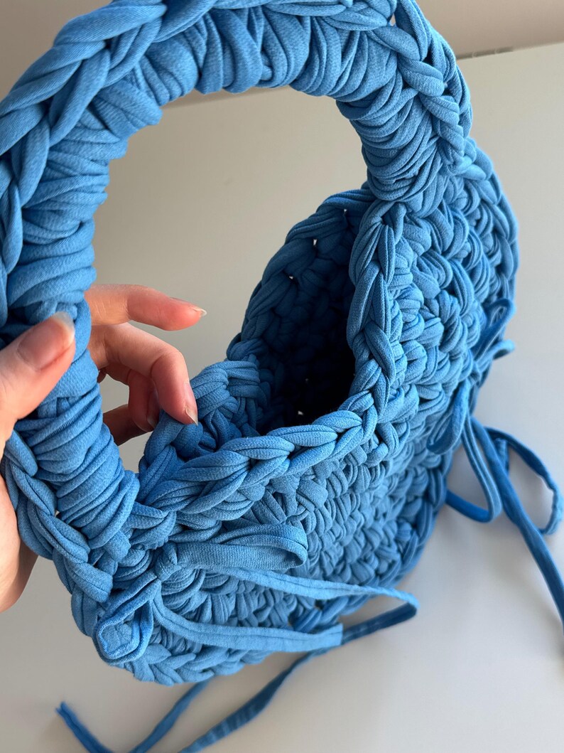 May include: A handmade, sky blue knitted handbag with a rounded handle. The bag is constructed from thick, braided fabric, and features a drawstring closure. The overall design is textured and soft, with a casual, yet stylish aesthetic.
