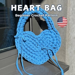 Può includere: Una borsa all'uncinetto a forma di cuore blu cielo con manico intrecciato e lacci lunghi. La borsa presenta il testo "HEART BAG" e "Beginner Crochet Pattern". Una bandiera americana è nell'immagine.