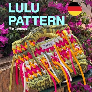 Op de afbeelding: Een handgemaakte gehaakte handtas met een levendig kleurenpalet van olijfgroene, felroze, oranje en witte stoffen stroken. De tas heeft geïntegreerde handvatten en een wit rechthoekig label met de zwarte tekst barwy aan de voorzijde.