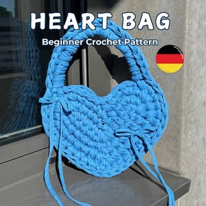 May include: A sky blue heart-shaped crochet bag with a braided handle and long ties. The text "HEART BAG" and "Beginner Crochet Pattern" are displayed on the image. The bag is made of thick, textured yarn.
