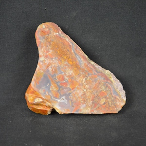 May include: A polished slab of agate stone featuring a triangular shape with a smooth, flat surface. The stone displays a complex pattern of swirling colours including burnt orange, deep red, creamy white, and translucent grey mineral inclusions.