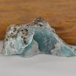 May include: A raw, natural gemstone with a striking blue hue, speckled with dark inclusions. The stone's rough texture and irregular shape are visible, set against a blurred wooden background. This unique mineral specimen is a beautiful example of natural artistry.